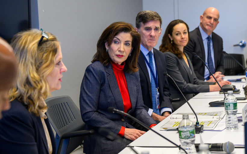 Governor Kathy Hochul, wearing a red turtleneck and blazer, sits at a table with other officials.