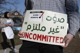 A sign in the foreground says "UNCOMMITTED" in red with Arabic text above, and a small image of a ballot with "uncommitted" checked and Biden unchecked. In the background is a woman in a hijab.