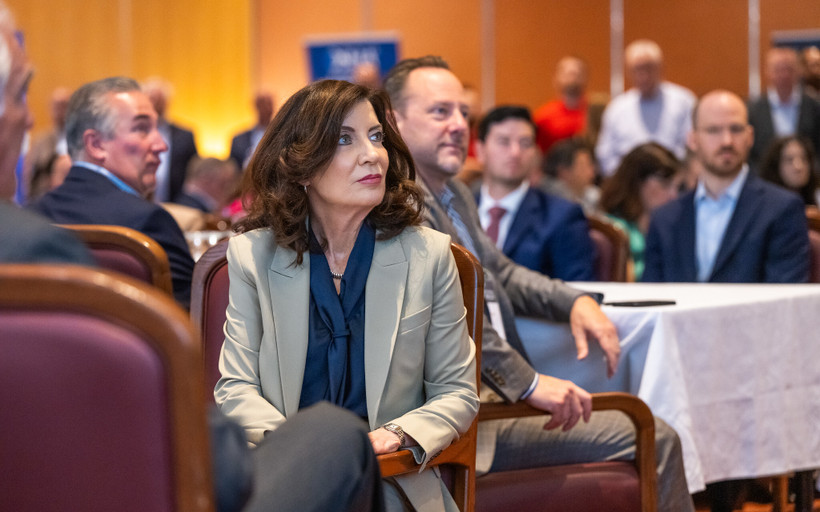 Governor Kathy Hochul sits in a chair at a breakfast meeting and looks up.