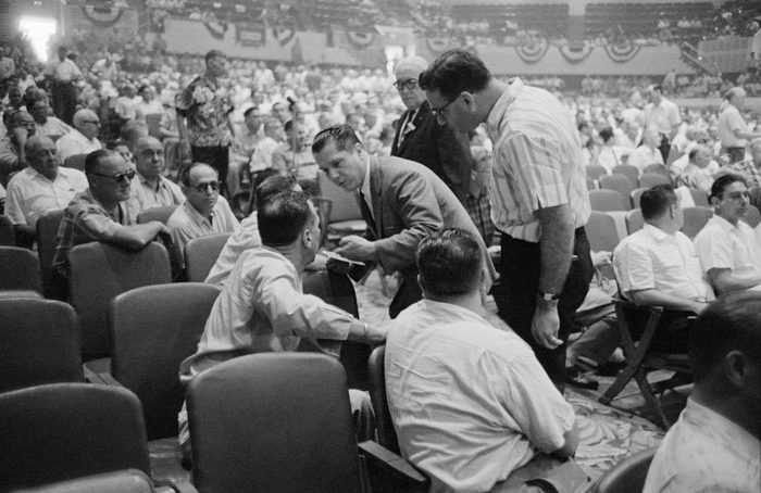 Men sit in a full auditorium. A man wearing a suit stands in the middle of the isle while leaning forward to talk to one of the men sitting down.