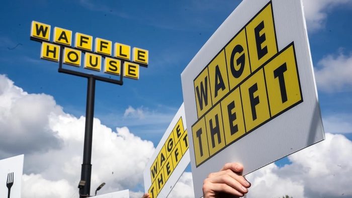 Below a Waffle House sign, hands hold "Wage Theft" signs against a blue sky