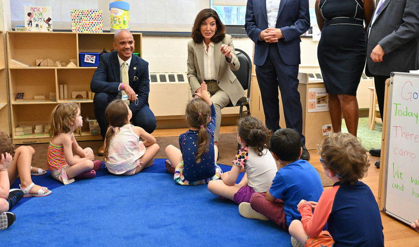 New York City Governor Kathy Hochul sits in front of a group of small children on a blue carpet.