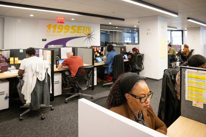 In an office, people work next to each other at desks with short walls in between them. The wall reads "1199 SEU."
