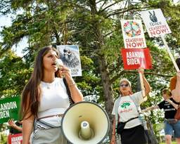 A woman protester shouts into a large megaphone in the foreground. A few older protesters stand behind her with signs reading "Gaza love," "Abandon Biden--ceasefire now" and have flower and peace sign imagery.