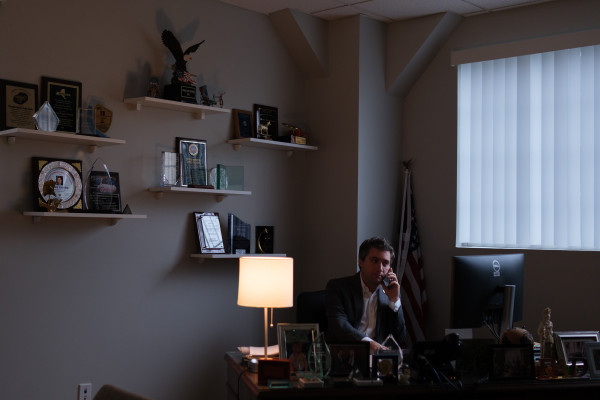 Senator James Skoufis sits at a large, dark desk illuminated by the soft glow of a lamp, speaking on the phone and looking away from the camera. Plaques and awards adorn the wall and desk.