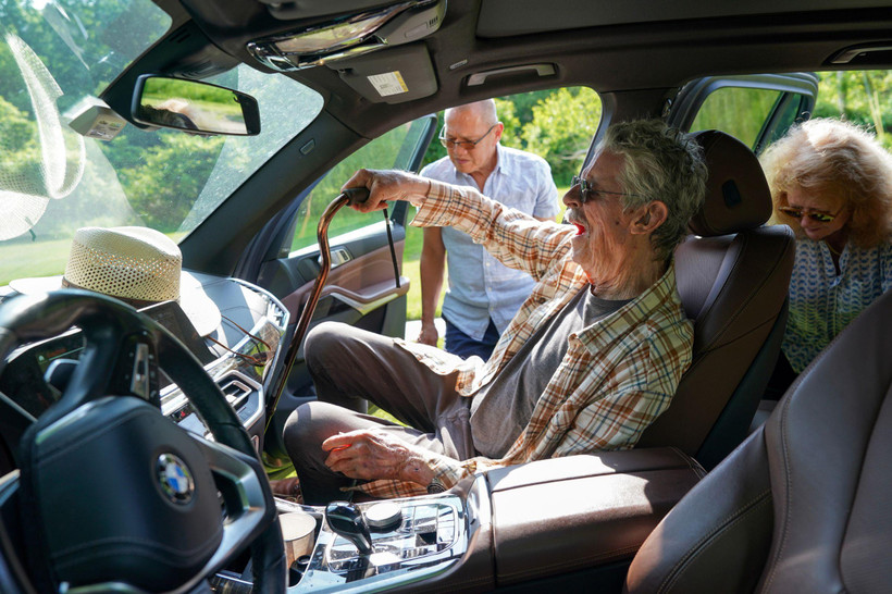 Gordon Chaplin sits in the passenger's seat of a car, holding a cane. A man on his right standing outside the car seems ready to assist him.