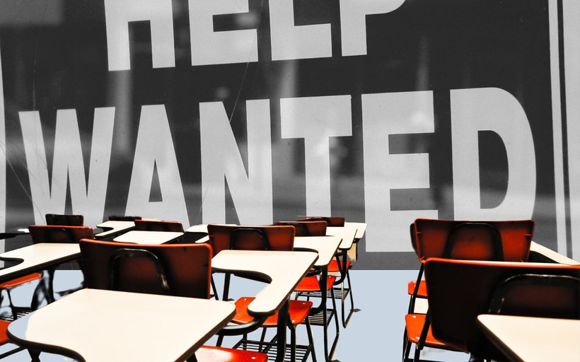 Empty schoolroom chairs face a large help wanted sign.