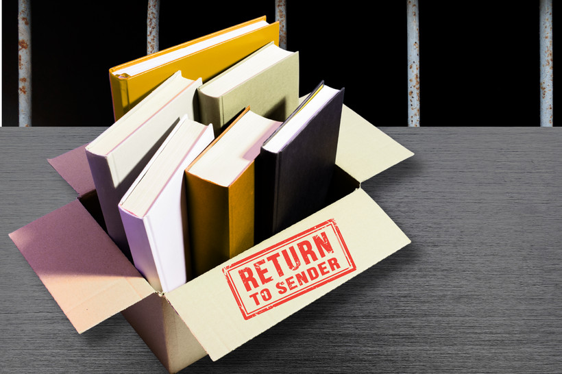 A box of books with a label reading "return to sender" sits on a metal table in front of prison bars.