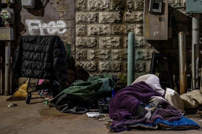 A person is bundled in a sleeping bag next to a pile of stuff. Graffiti on a stone wall behind them reads "Free."