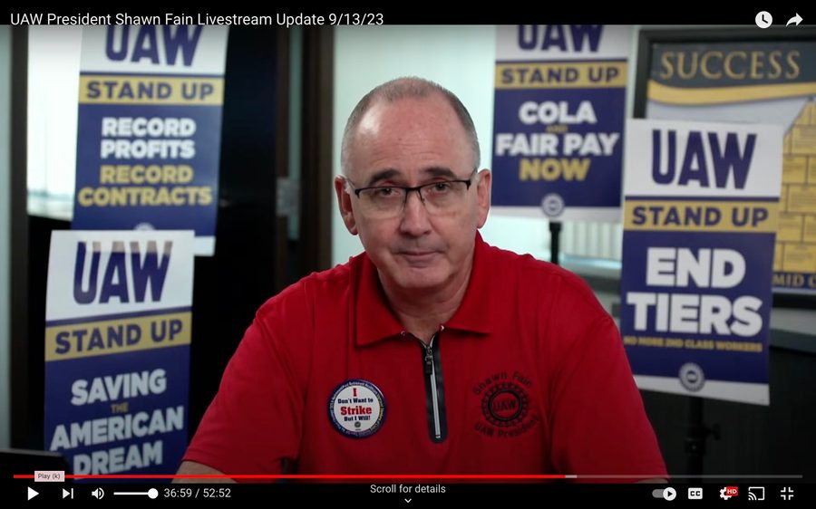 Screenshot of United Auto Workers President Shawn Fain in a livestream wearing a red UAW shirt, a UAW pin, and surrounded by UAW signs with demands like ?"End Tiers" and "COLA Fair Pay"