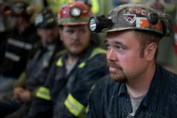 A row of coal miners sit with lanterns on their helmets. The front one, in focus, looks worried.