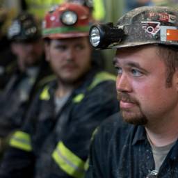 A row of coal miners sit with lanterns on their helmets. The front one, in focus, looks worried.