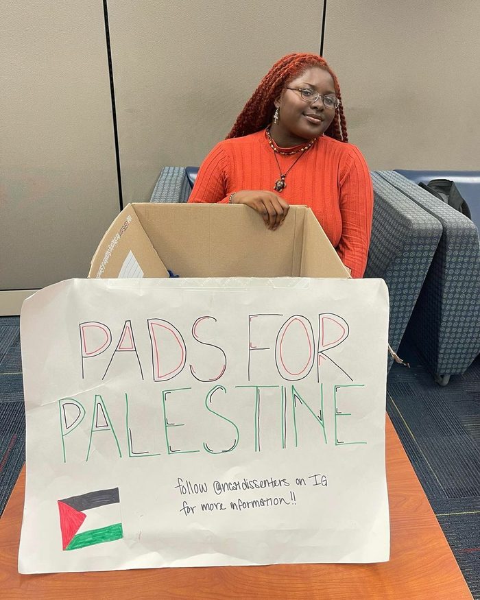 Smiling student with braids and orange shirt sits behind a giant box labelled "PADS FOR PALESTINE"