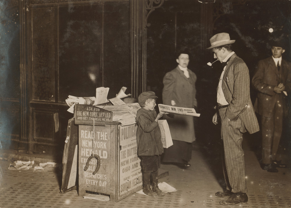 A sepia toned photograph of a child offering a newspaper to a man in a hat with a pipe.