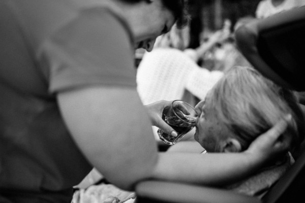 A standing woman wearing scrubs gently cradles a seated older woman's head while she helps her drink from a glass.