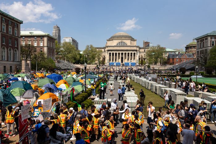 In an expansive view of the campus center at Columbia University, a crowd of people wear orange and yellow safety vests in the foreground, with an encampment of tents to the left.