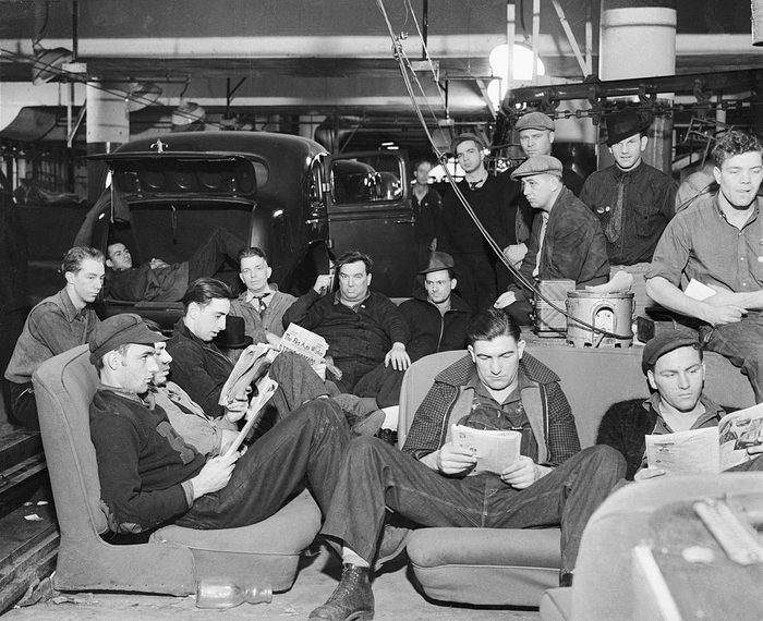 Black-and-white photo: Men sit reading in chairs with old-fashioned cars behind them