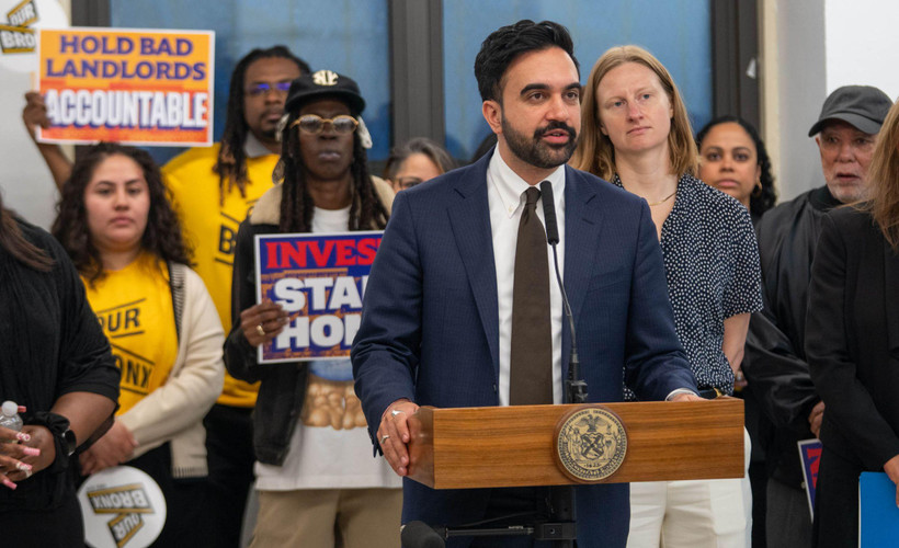 At a press conference, Mayor Zohran Mamdani stands speaking at a podium, behind and to his left stands Cea Weaver, director of the New York City Mayor's Office to Protect Tenants. Behind them stand people holding signs bearing the words "Hold bad landlords accountable" and "Invest in stables homes."
