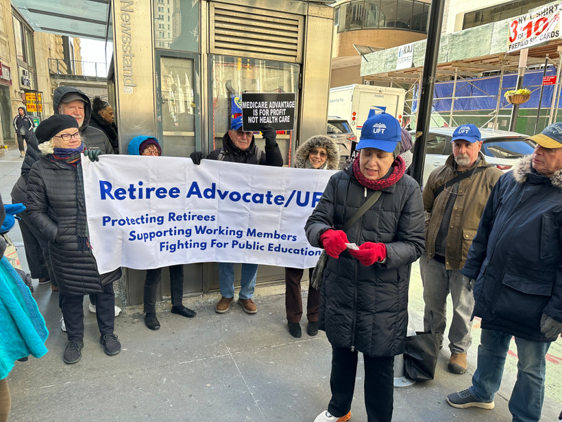 People stand in front of a New York City newsstand holding a sign that says "Retiree Advocate/UFT Protecting Retirees Supporting Working Members Fighting for Public Education"