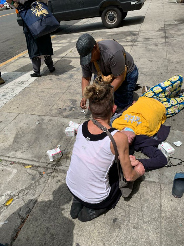 Someone lays on the ground, surrounded by two people that are checking his pulse and helping administer the Narcan nasal spray.