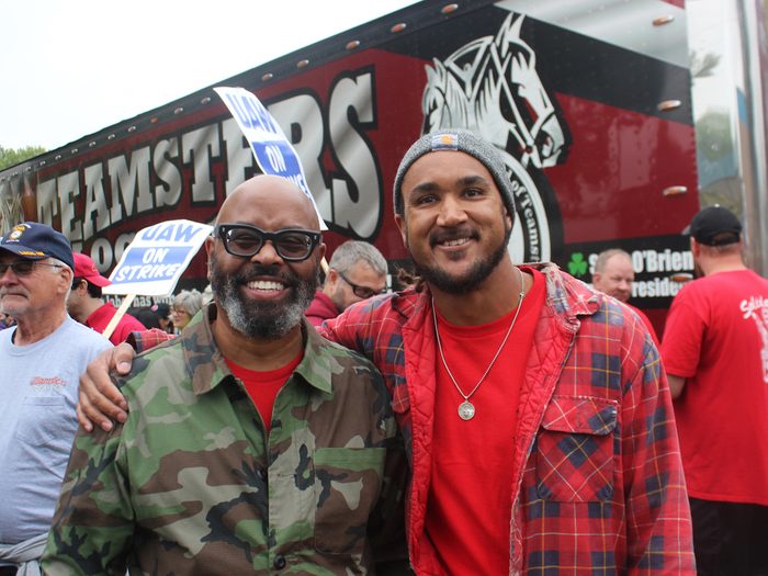 A younger man in red stands with his arm around a man in glasses, a camo jacket and a partially gray beard. They are smiling, and behind them are protests signs and Teamsters truck.