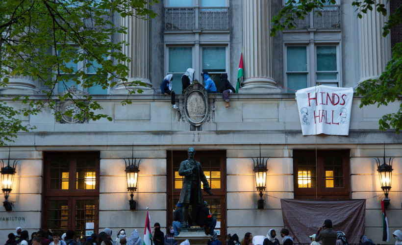 Student protesters gather at Columbia's Hamilton Hall or "Hind's Hall," renamed after Hind Rajab, a six-year-old Palestinian girl who was shot to death with paramedics on an IDF-supplied emergency route. On the right, they hang a banner that says "HIND'S HALL." Protesters sit up high on a facade.