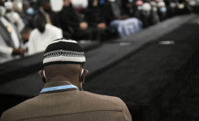 Close-up photo of a man from behind wearing a suit, kufi, and mask. In the background, many other people attending a funeral.