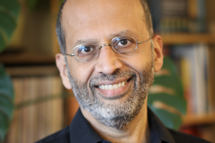 Deepak Bhargava smiles while wearing a black button-up and rectangular wire-framed glasses. A monstera plant and book shelves are in the background. He has a salt-and-pepper beard.
