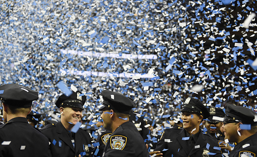 New NYPD officers smile amid blue and white confetti at police academy graduation ceremony.