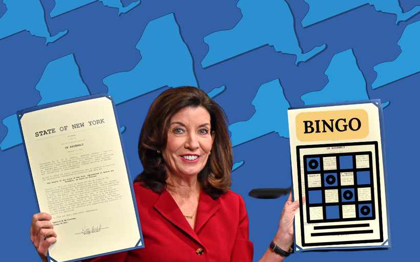 Kathy Hochul holding up papers in each hand. One hand has an official State of New York document and the other is a Bingo game card that was illustrated.