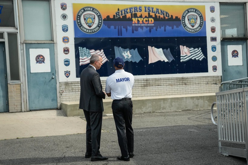 Mayor Eric Adams standing with his back to the camera, facing a poster reading "Rikers Island NYCD".