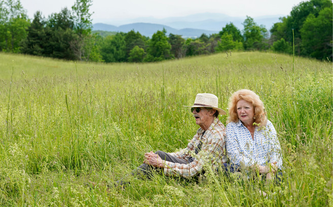 A man and woman sit in a field of tall grass. In the distance, trees and mountains are visible.