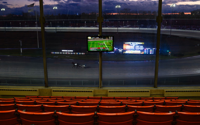 Rows of empty seats watch a horse racing track with a lone harness racer and horse.