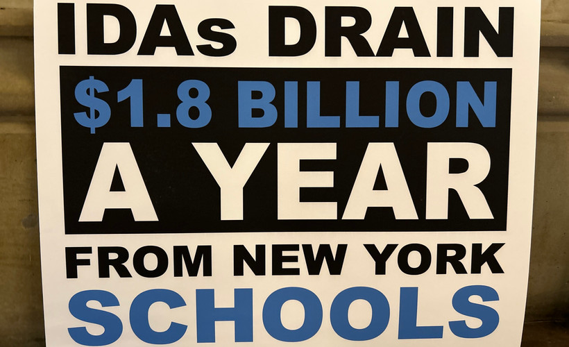 A sign reads "IDAs DRAIN $1.8 BILLION A YEAR FROM NEW YORK SCHOOLS."