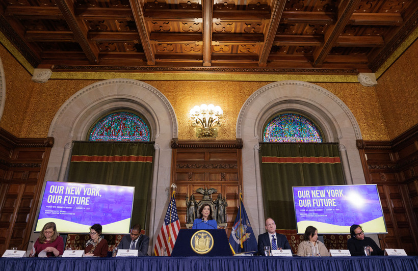 Governor Kathy Hochul stands at a podium, flanked by staff, in a room with two high, arched windows and two screens that read "Our New York, Our Future."