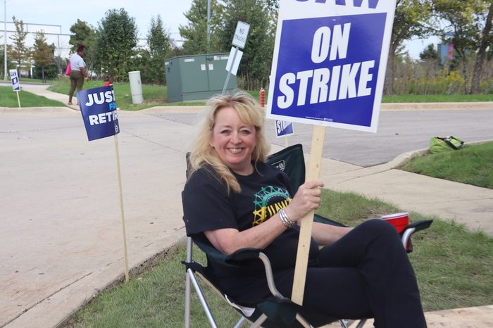 A woman in all black sitting in a chair with a "UAW on Strike" sign