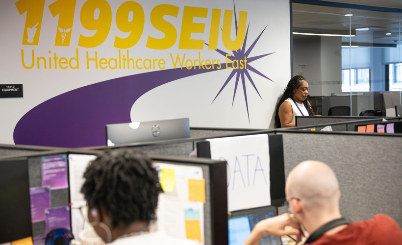 In the 1199 SEIU office, people work in at desks with a large 1199 SEIU: United Healthcare Workers East logo on the wall.