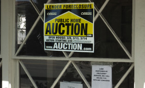 Window looking into a darkened foreclosed home. A yellow sign says "LENDER FORECLOSURE" and "PUBLIC HOME AUCTION." Smaller paper signs say "Warning No Trespassing" and note a lawn maintenance company.