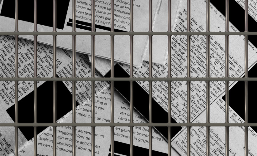 a stack of newspapers, with redaction boxes covering the text, behind prison bars