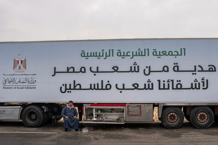 A large truck with Arabic writing across it sits parked, with the driver sitting in front of it.