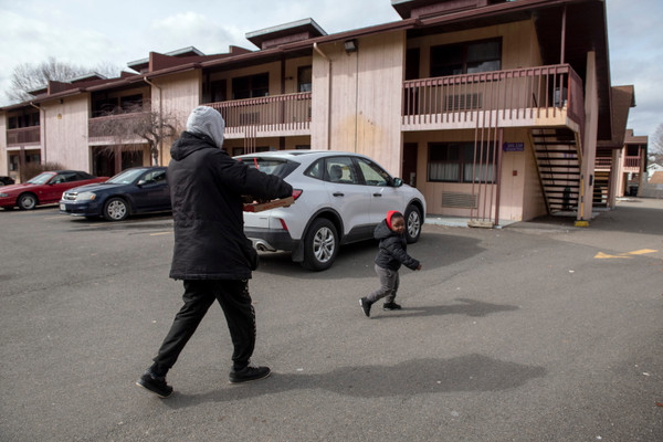 An adult and child wearing winter coats walk across a motel parking lot with three cars parked in it. The child is smiling and turns back to look at the adult, who is facing away from the camera.