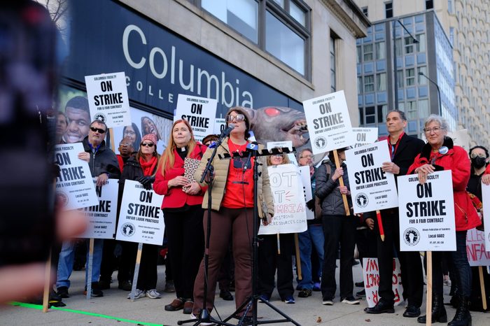 In front of Columbia College, a person wearing a beanie, sunglasses, and a coat stands behind microphones, flanked by a group of union members holding picket signs, most of which read "Columbia Faculty On Strike for a Fair Contract."