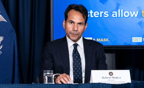 Former state budget director Robert Mujica sits at a table with a name card wearing a suit.