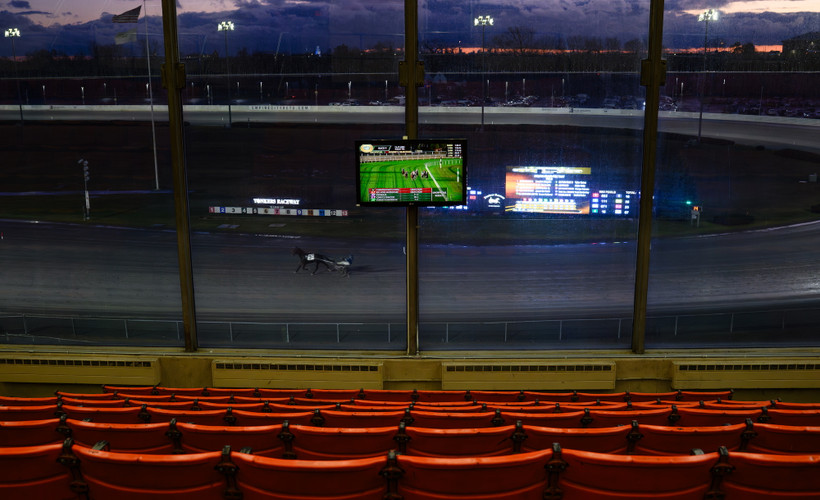 Rows of empty seats watch a horse racing track with a lone harness racer and horse.