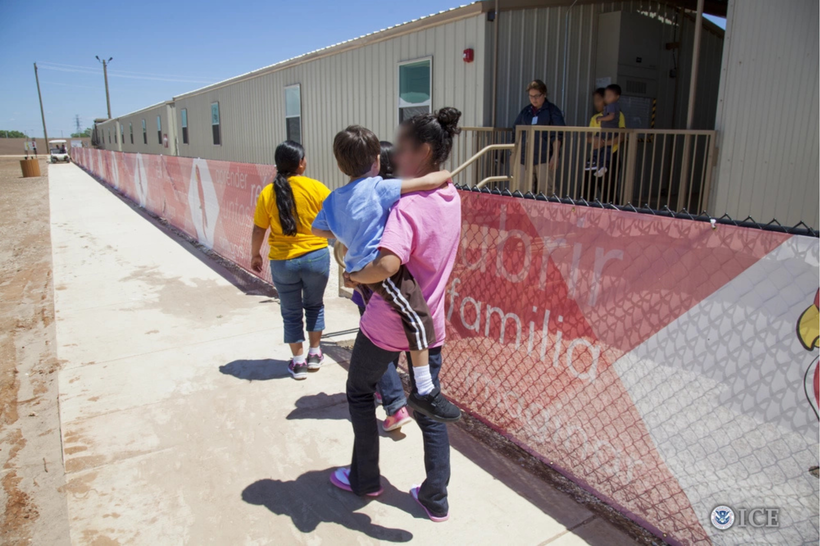 Three people, one of them holding a child, walk on a sidewalk near a prefabricated building in a desert-like area.
