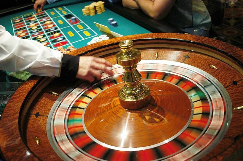 Closeup of a hand spinning a roulette wheel, with hands moving betting tokens in the background.