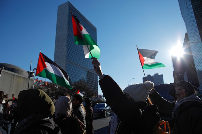 People in coats and jackets extend Palestinian flags into the air while walking across a street as the sun glints off of buildings in the distance.