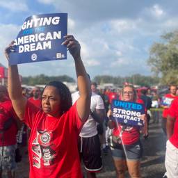 Photograph of a worker in a red shirt holding a sign that says "Fighting for the American Dream," with other marching behind her.