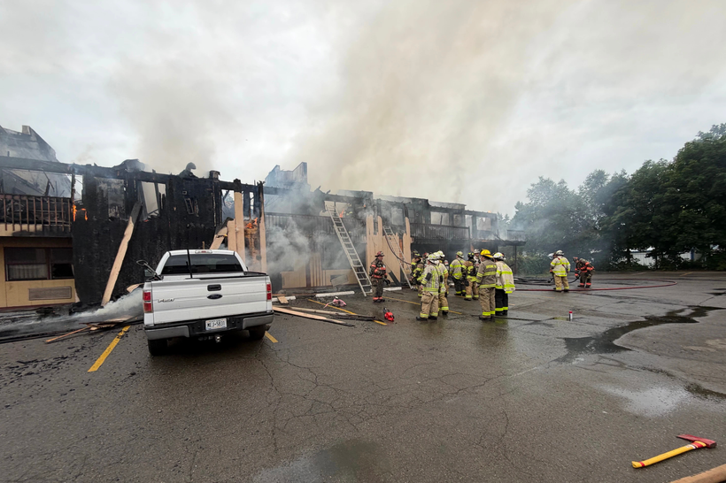 Firefighters stand next to a motel, which is pouring smoke with small mostly extinguished flames. Large parts of the motel are completely burned through, and buildings and sky are visible behind it.