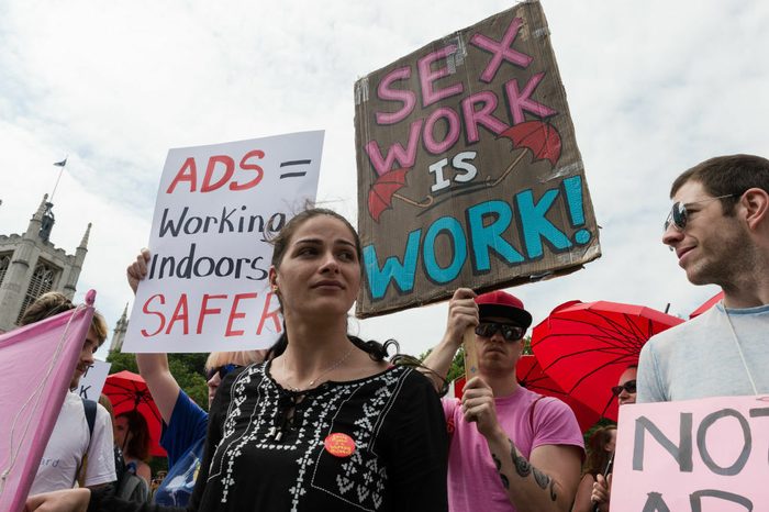 A woman looks out with signs behind her saying "Ads = Working Indoors = Safer" and "Sex Work is Work"